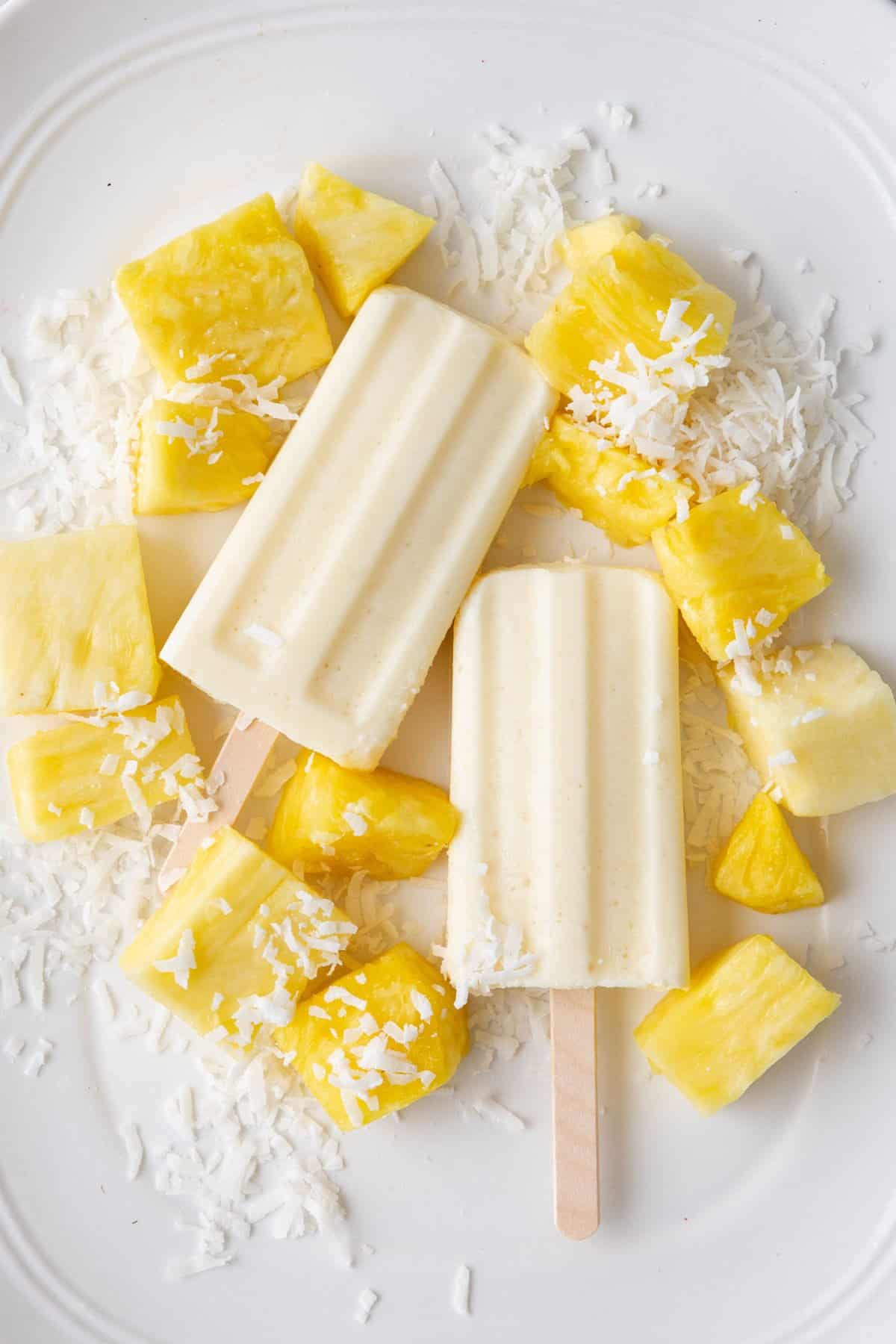 Frozen pineapple coconut cottage cheese pops on a large platter with fresh pineapple chunks and shredded coconut around.