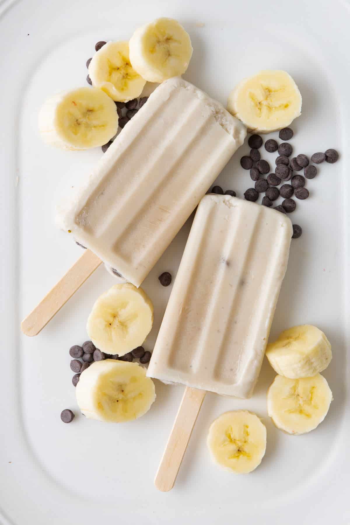 Frozen Banoffee pie popsicles on a large platter with sliced bananas and chocolate chips around.