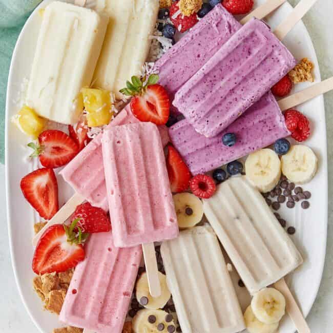 Frozen cottage cheese ice cream popsicles on a large tray: strawberry cheesecake, banoffee pie, pineapple coconut, and mixed berry breakfast pops; extra berries, granola, chocolate chips, and shredded coconut around.