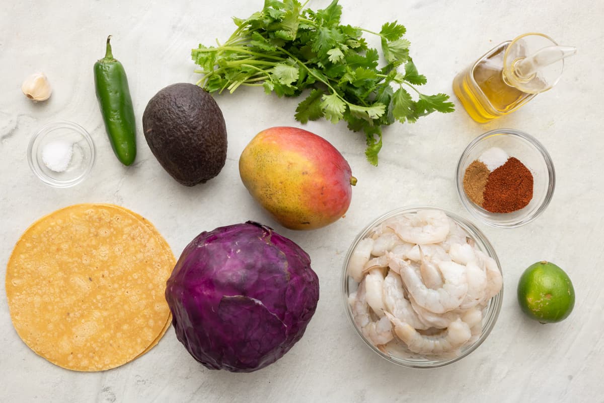 Ingredients for recipe: garlic, salt, jalapeño, avocado, corn shells, head of red cabbage, mango, cilantro, oil, spices, raw shrimp, and lime.