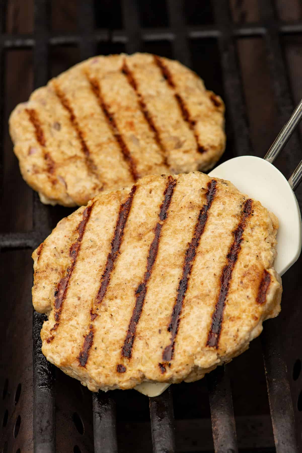 Yogurt chicken burger patties on a grill with a spatula lifting one up to show grill marks.