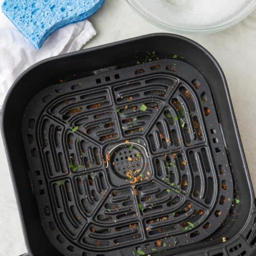 A dirty air fryer basket with cleaning supplies and soapy water nearby.