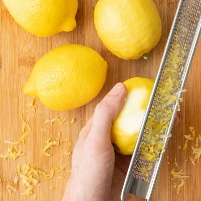 Lemons on a cutting board with a hand holding one while using a microplane to zest it.