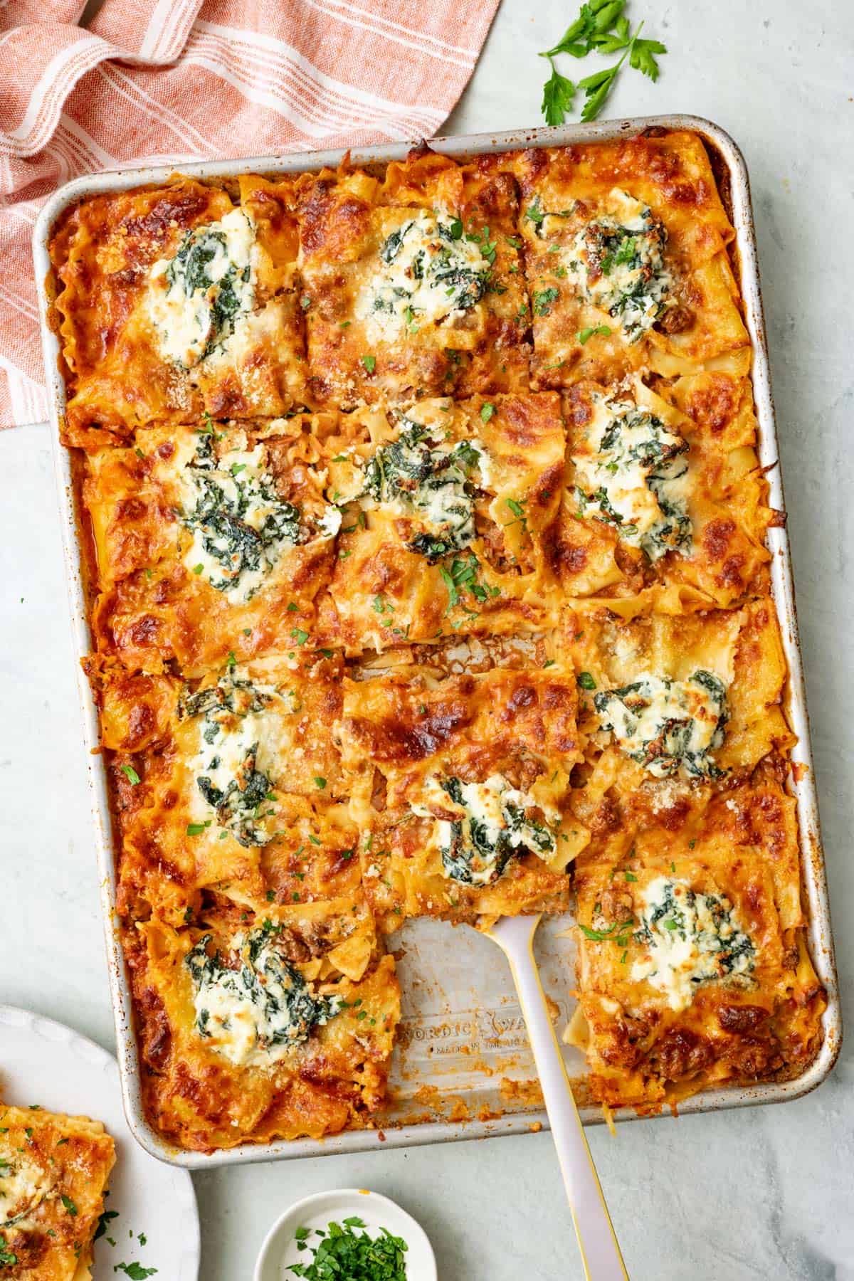 Lasagna cut into 12 pieces with one sliced removed onto a plate to the side and a spatula lifting up another serving.