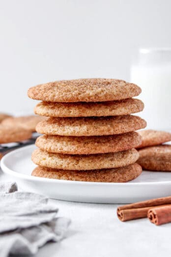 Snickerdoodles stack tall on a round plate with extra cookies nearby and a glass of milk in the background.