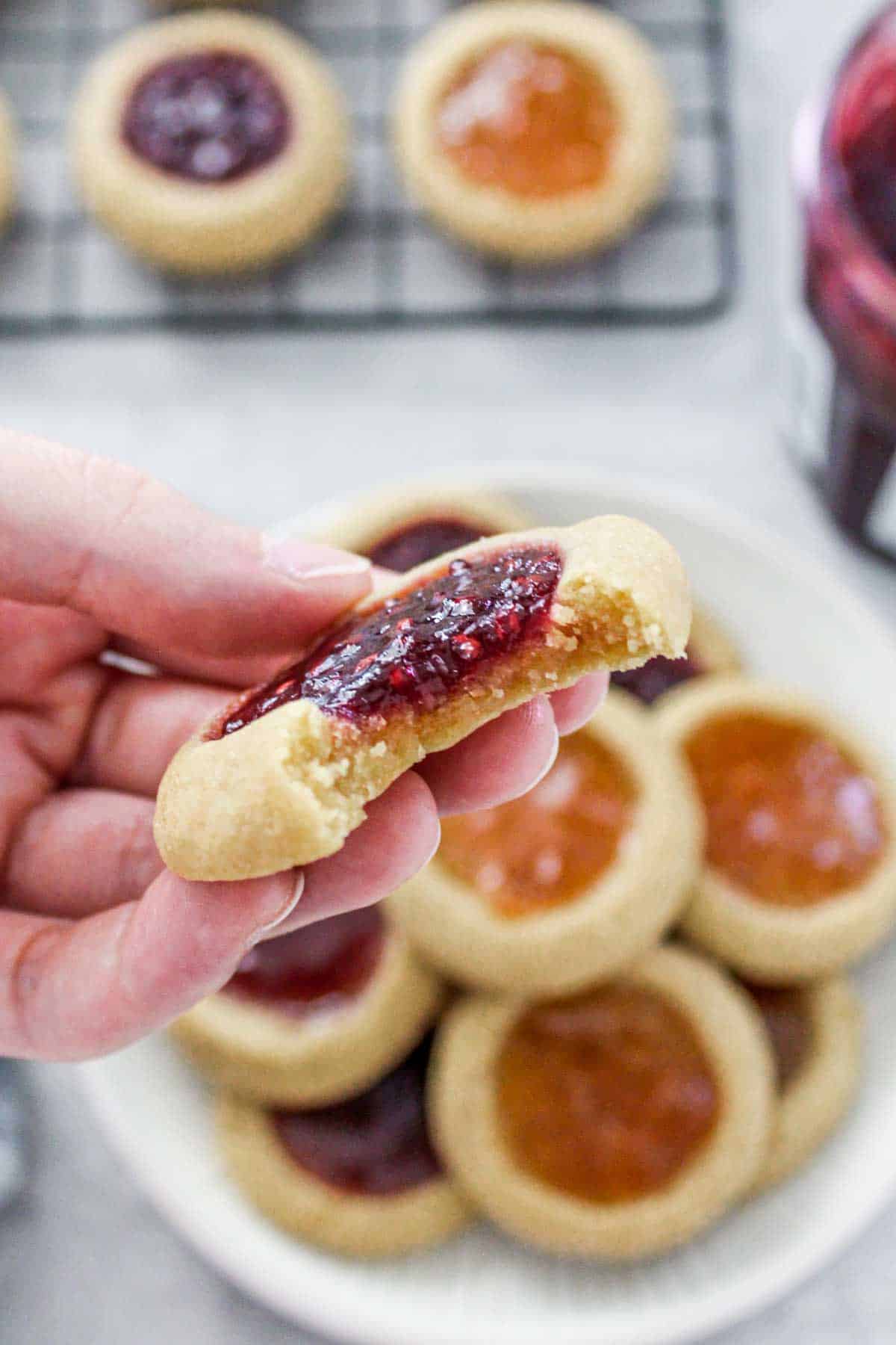 Hand holding one thumbprint cookie with a bite taken with the plate and rack of more cookies below.