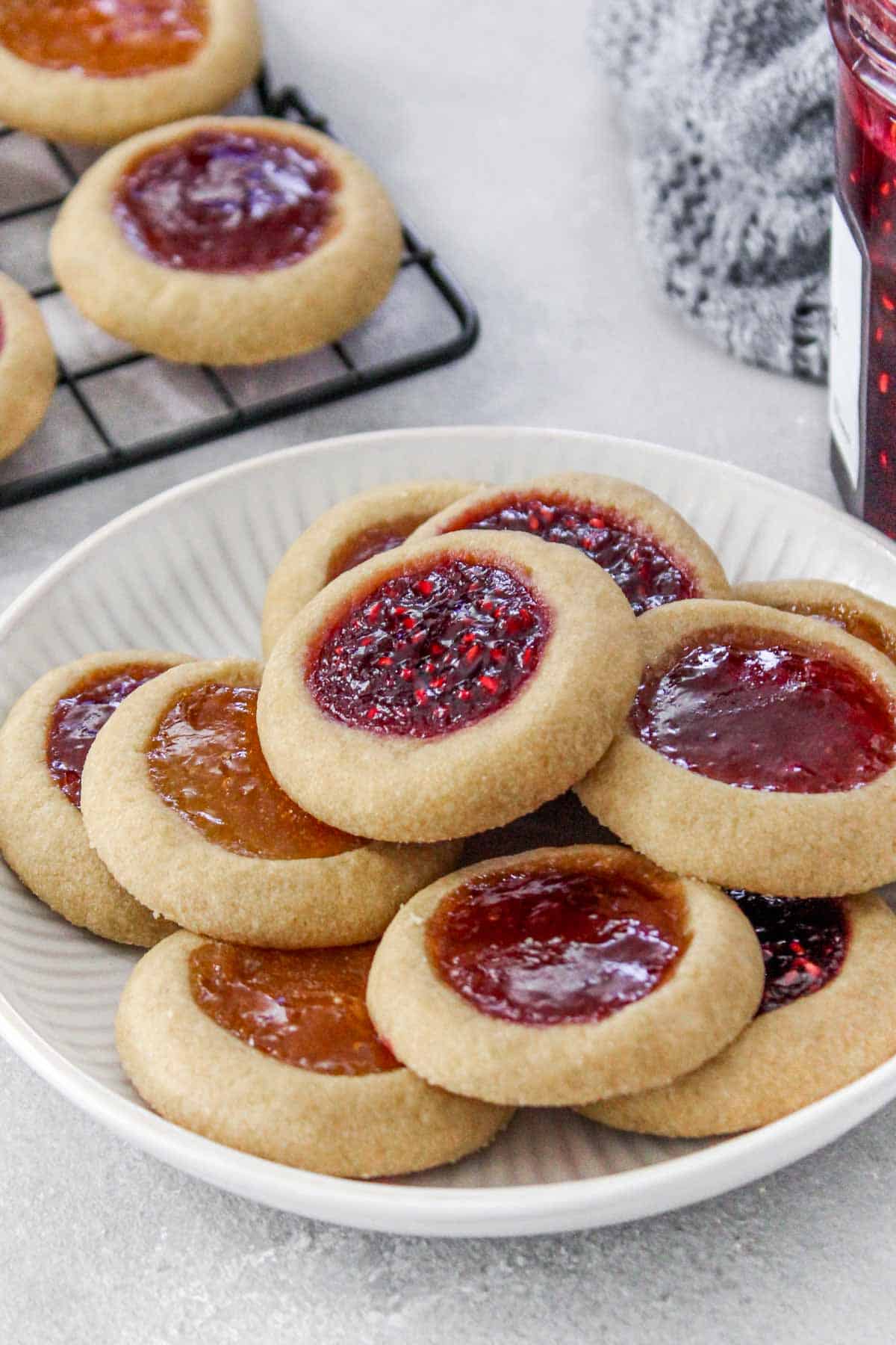 Thumbprint cookies on a plate with a wire rack of more cookies and a jar of jam nearby.