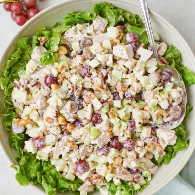 Waldorf salad on top of leafy lettuce in a large shallow bowl with a spoon dipped in and extra grapes and walnuts nearby.