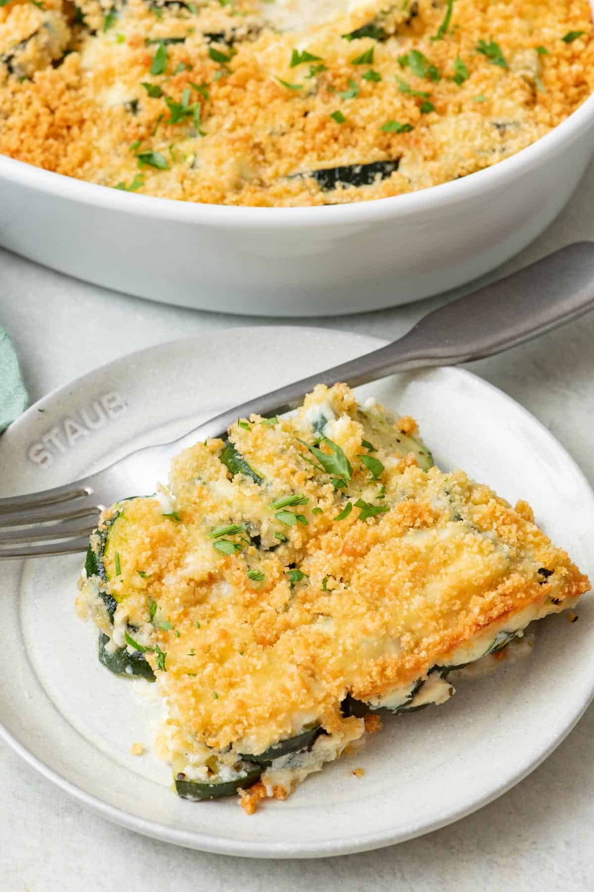 Zucchini casserole with a golden brown top on a small plate with a fork and the casserole dish nearby.