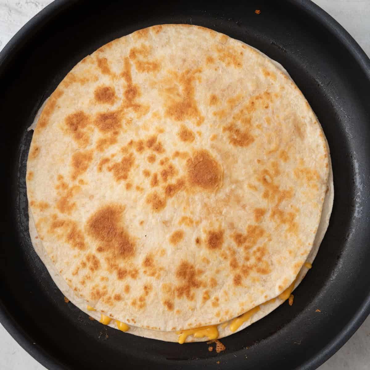 Tortilla flipped in pan to show golden brown marks and melted cheese from side.