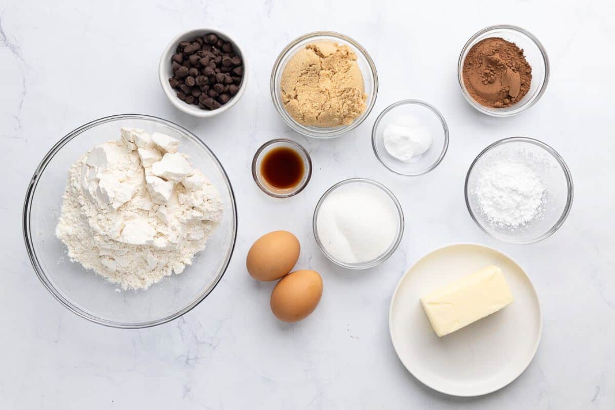 Ingredients for recipe in individual bowls: flour, chocolate chips, brown sugar, vanilla, eggs, sugar, rising agents, cocoa powder, and butter.