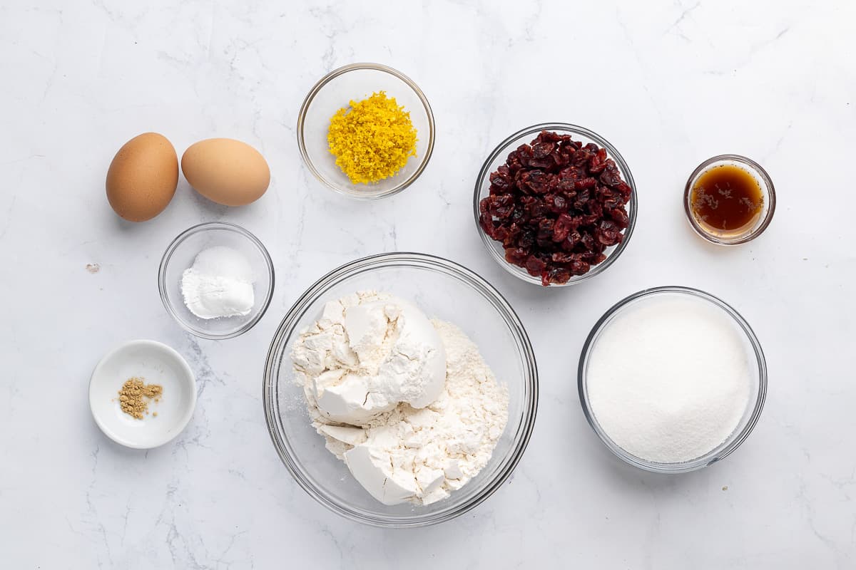 Ingredients for recipes in individual bowls: eggs, rising agents, ginger, flour, cranberries, vanilla, and sugar.