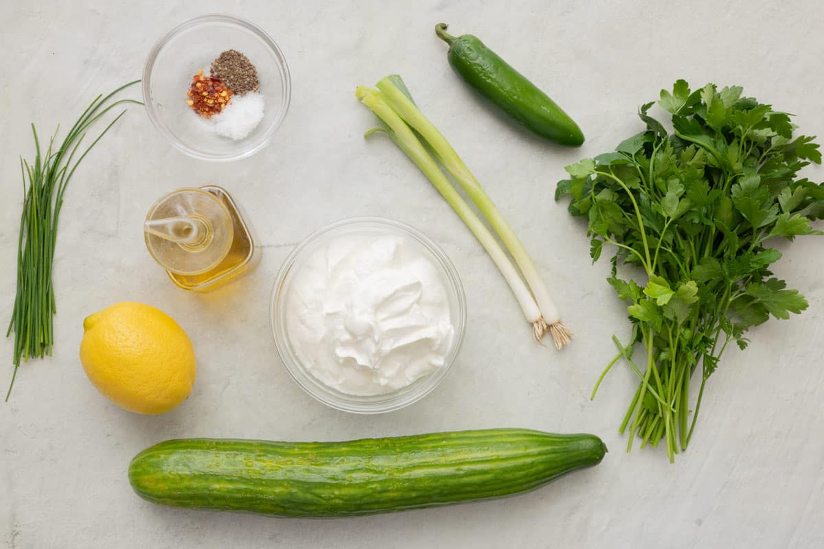 Ingredients for recipe before prepping: chives, salt, pepper, and red pepper flakes, oil, lemon, cucumber, yogurt, green onion, jalapeno, and fresh parsley.