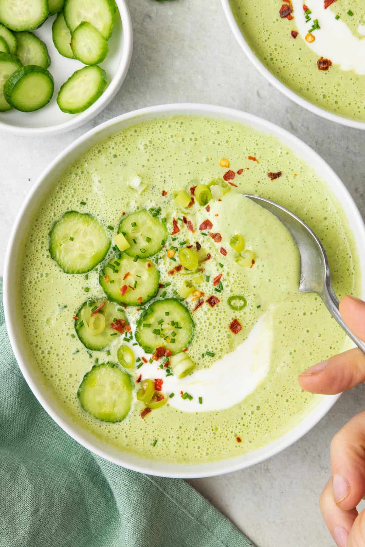 Cucumber gazpacho garnished with cucumber slices, yogurt, red pepper flakes, and green onions with a spoon dipping into the bowl.