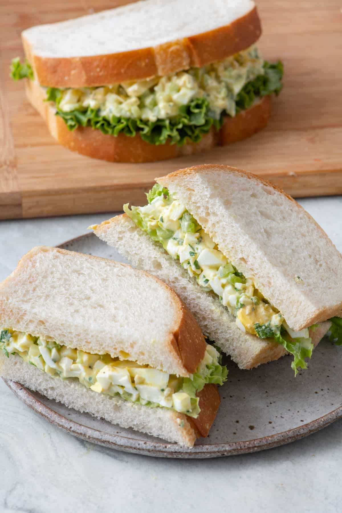 Egg salad sandwich cut in half on a plate with a whole sandwich nearby.