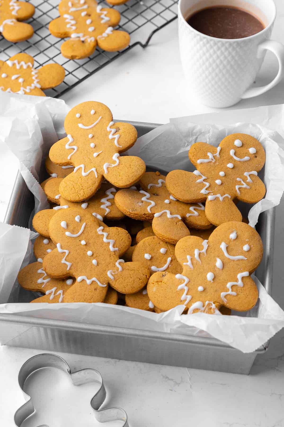 Gingerbread cookies in a a parchment lined square tin decorated with icing. Gingerbread man cookie cutter nearby and a wire rack with more cookies and a cup of coffee, too.