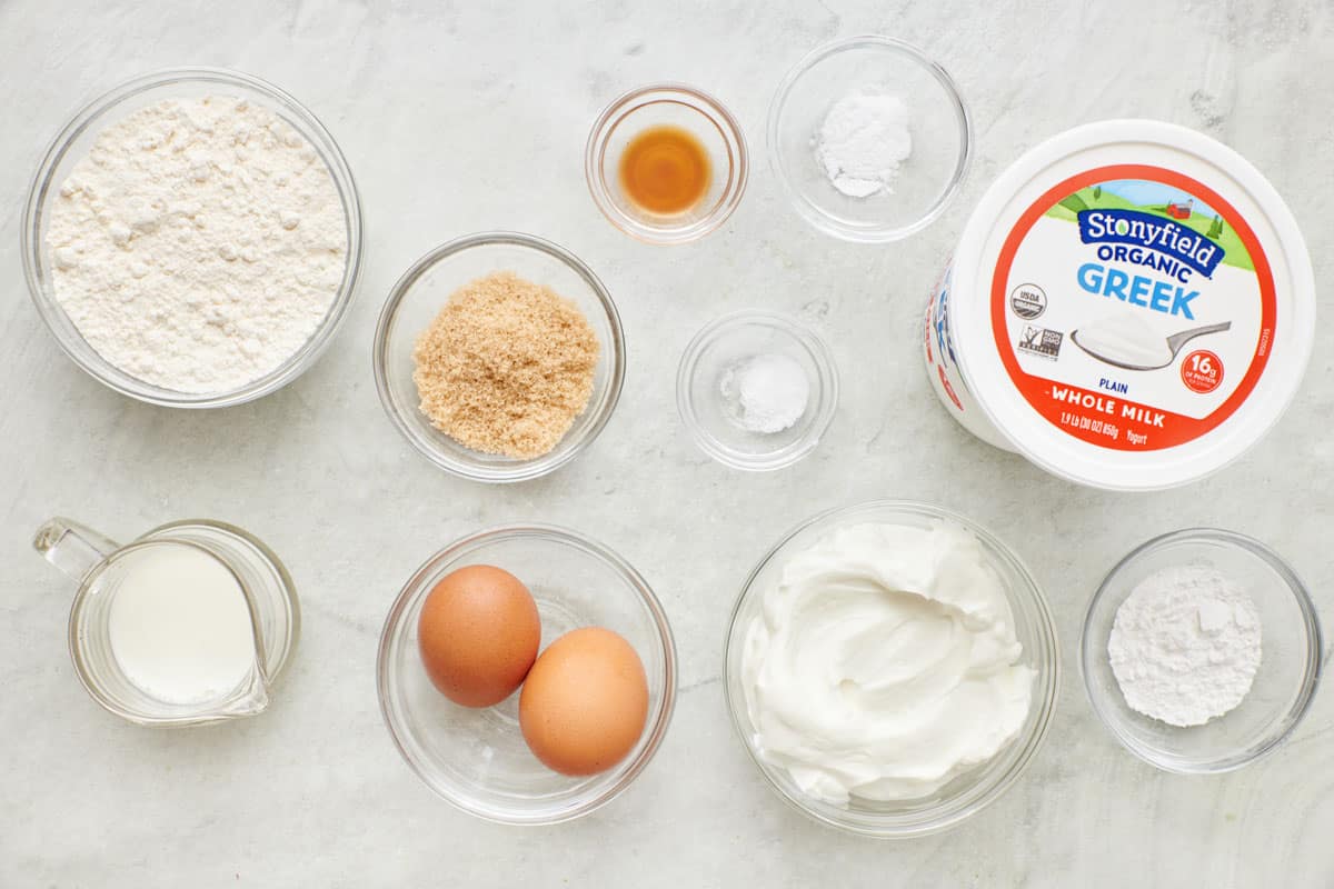 Ingredients for recipe in individual bowls: flour, milk, brown sugar, eggs, vanilla, rising agents, salt, Stonyfield Organic Greek Yogurt in a bowl with tub nearby.