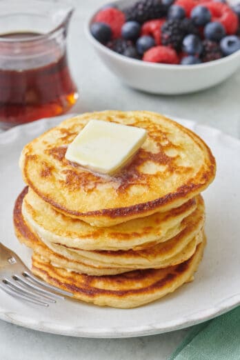 Stack of Greek yogurt pancakes with a pat of slightly melted butter on top and a small jar of syrup and fresh berries nearby.
