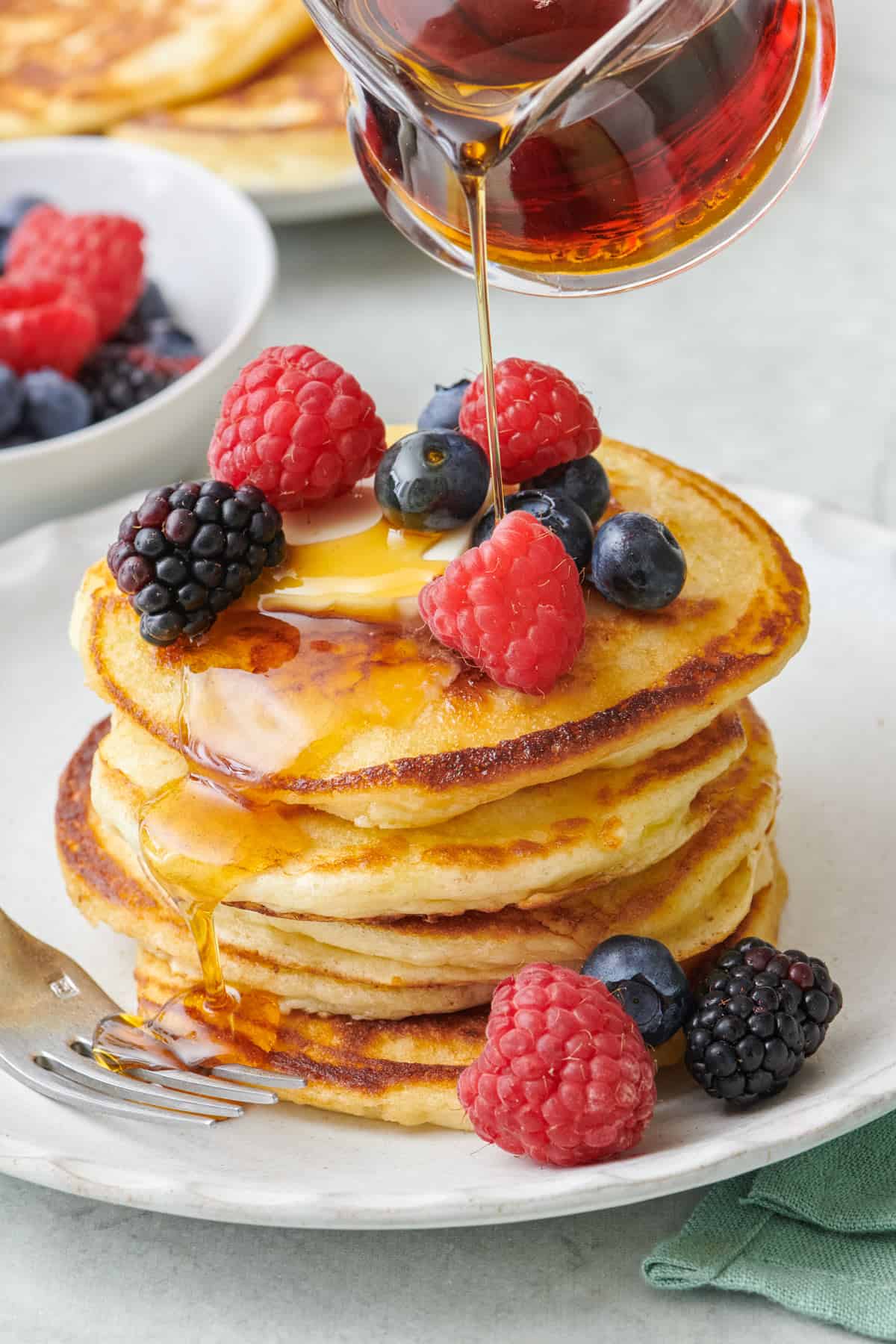 Pouring maple syrup on top of a large stack of Greek yogurt pancakes topped with fresh mixed berries.