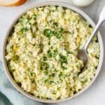 Egg salad in a large shallow serving bowl with spoon dipped in, garnished with fresh parsley and black pepper.