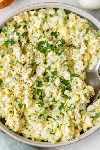 Egg salad in a large shallow serving bowl with spoon dipped in, garnished with fresh parsley and black pepper.