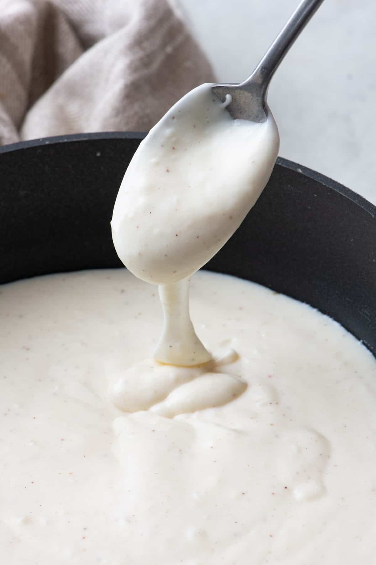 Spoon lifting up bechamel sauce as it drizzles back into pot to show thickened consistency.