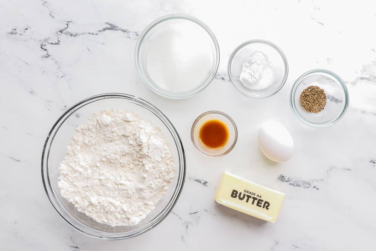Ingredients for recipe before prepping in individual bowls: flour, sugar, rising agents, anise seeds, vanilla, an egg, and butter stick.