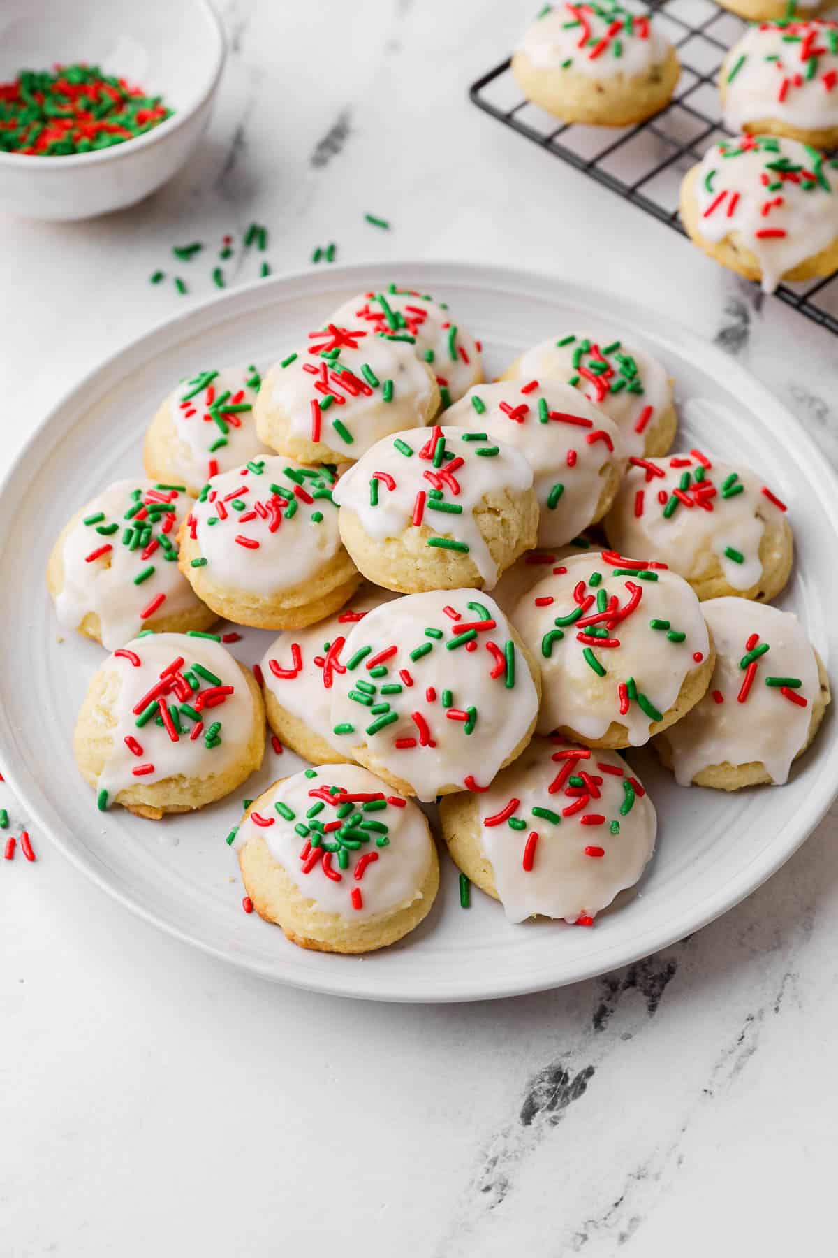 Italian iced christmas cookies with decorated with holiday green and red sprinkles with a small bowl of extra sprinkles nearby and a cooling rack with more cookies.