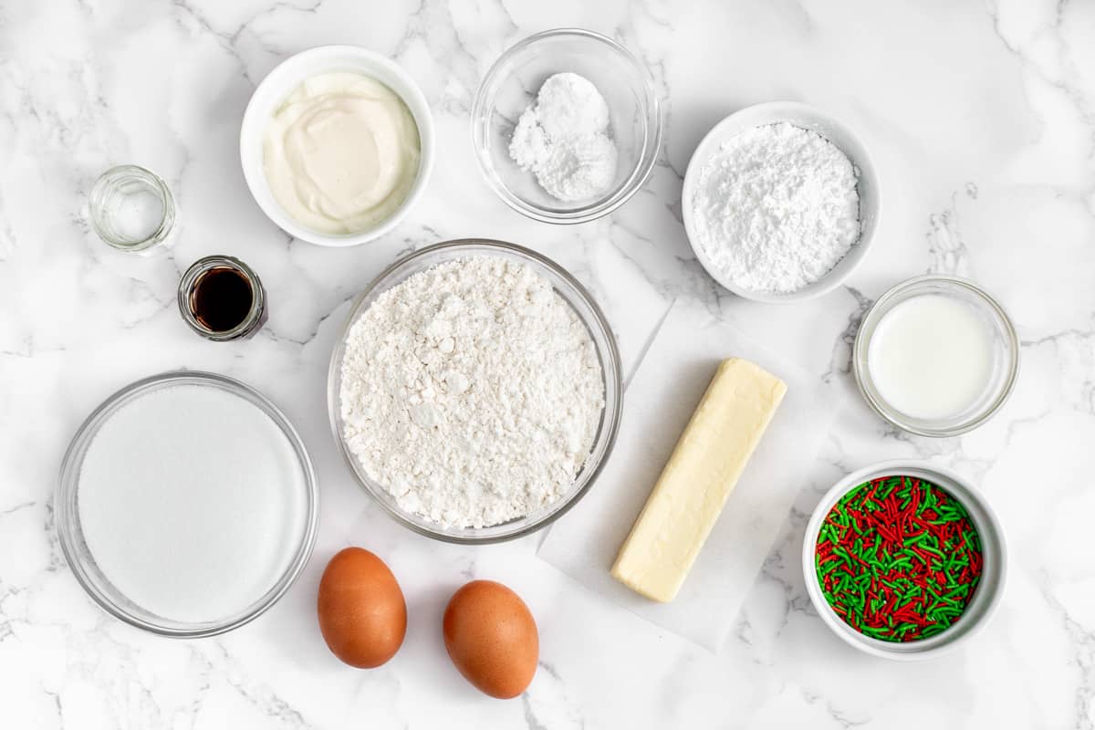 Ingredients for recipe in individual bowls: almond extract, vanilla extract, sugar, greek yogurt, flour, eggs, salt and rising agents, powdered sugar, stick of butter, milk, and holiday sprinkles.
