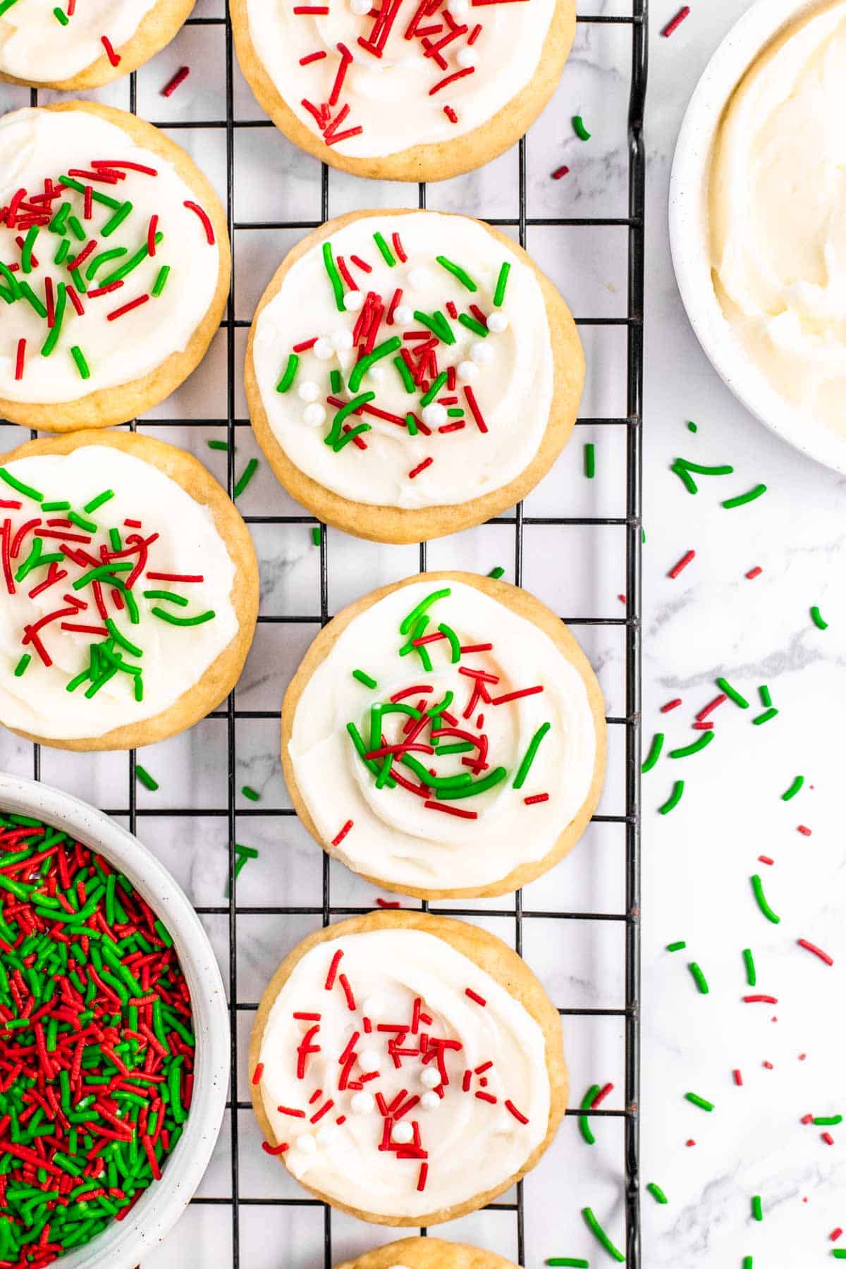 Close up of Lofthouse cookies on a wire rack decorated with holiday sprinkles with an extra bowl of sprinkles on the rack and frosting nearby.