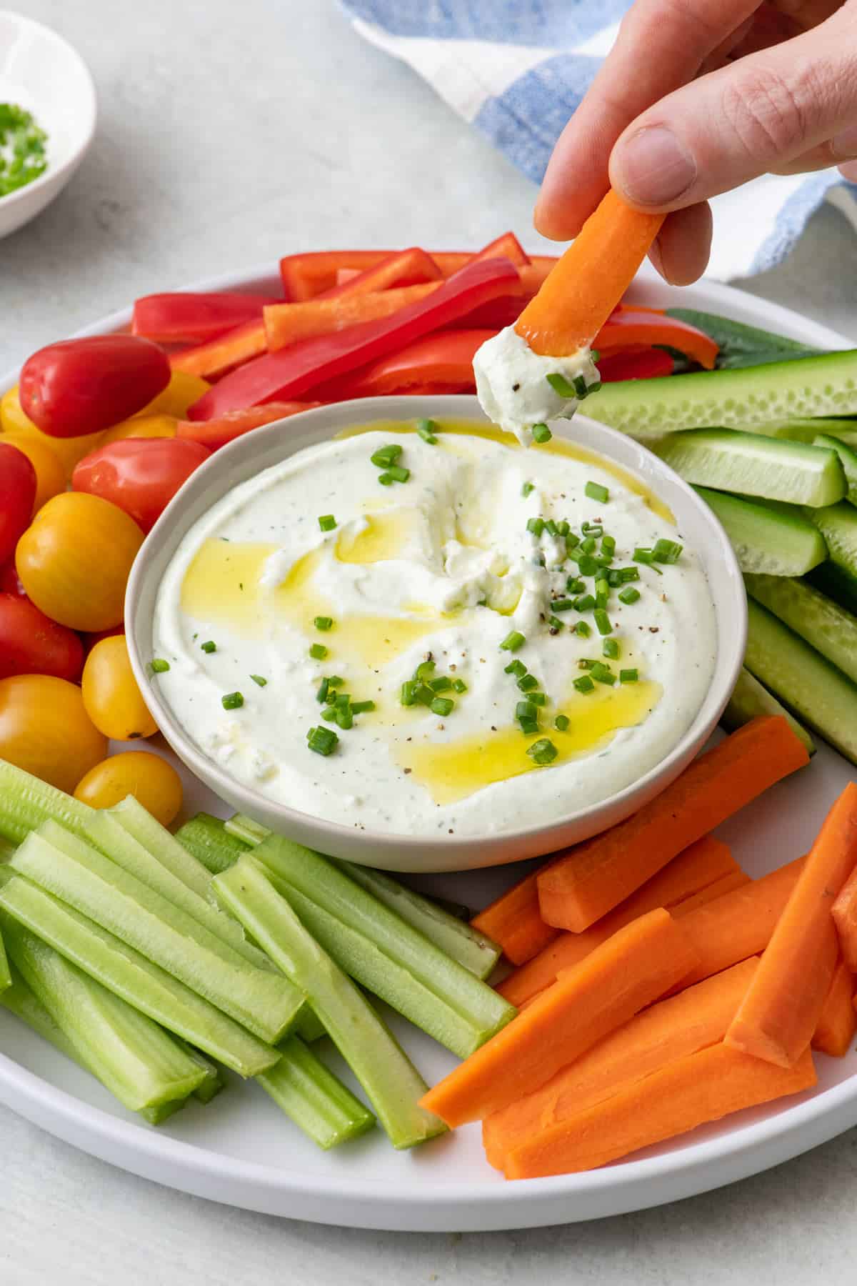 Carrot being dipped into a bowl of whipped cottage cheese garnished with chives and olive oil. Bowl on a platter surrounded by more fresh veggies cut into sticks.