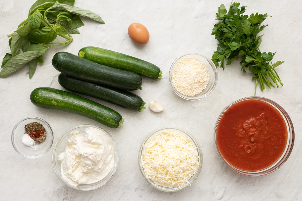 Ingredients for recipe before prepping: basil, zucchini, egg, seasonings, garlic clove, breadcrumbs, grated parmesan, ricotta, marinara sauce, and fresh parsley.