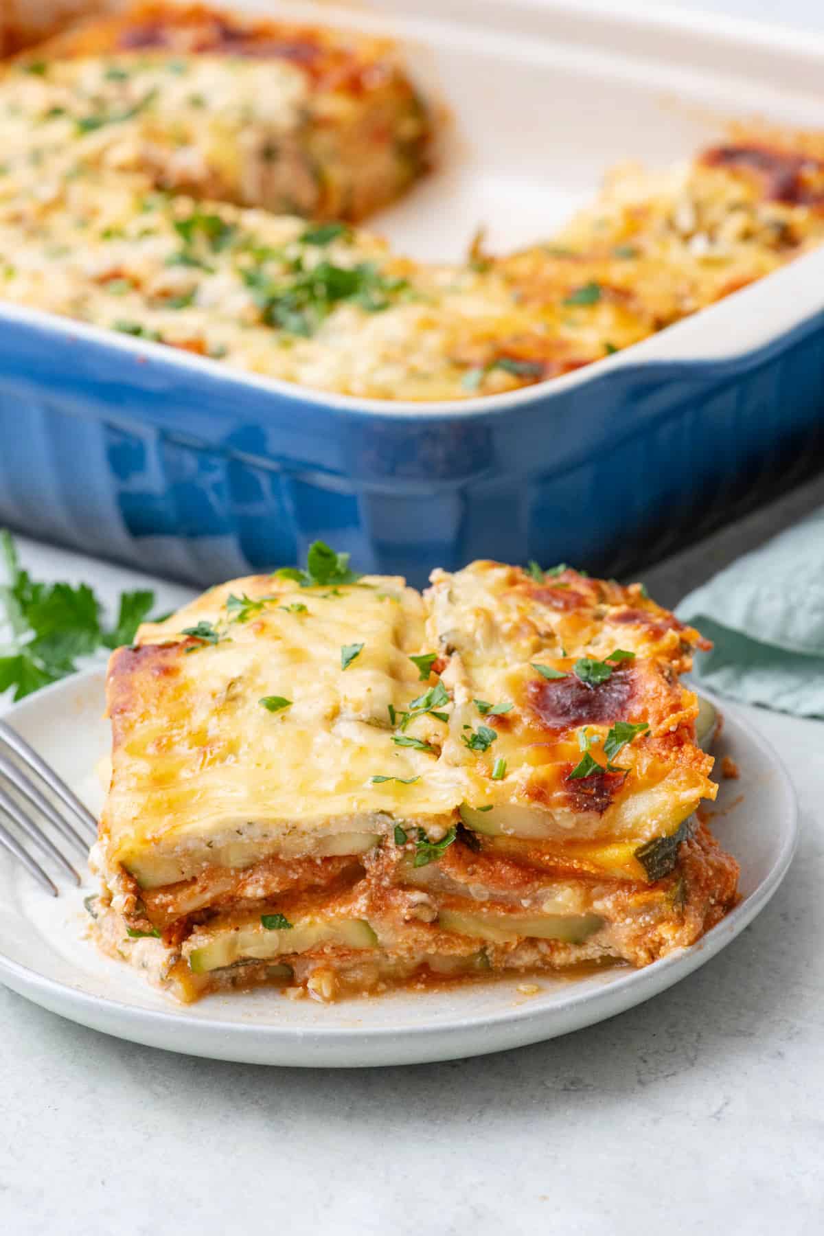 Serving of zucchini lasagna on a small plate to show layers with baking dish of lasagna behind plate showing a serving removed.