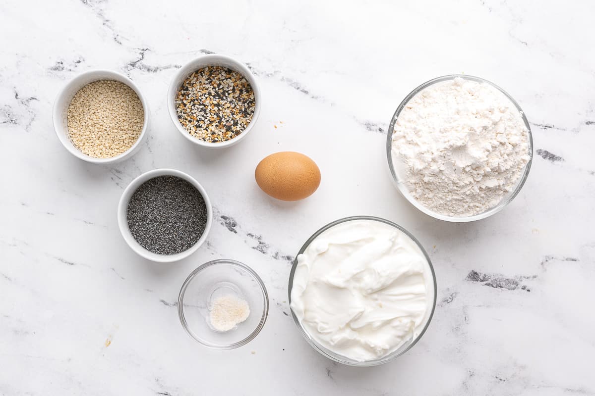Ingredients for recipe in individual bowls: sesame seeds, everything but the bagel seasoning, poppyseeds, salt, an egg, Greek yogurt, and self-rising flour.