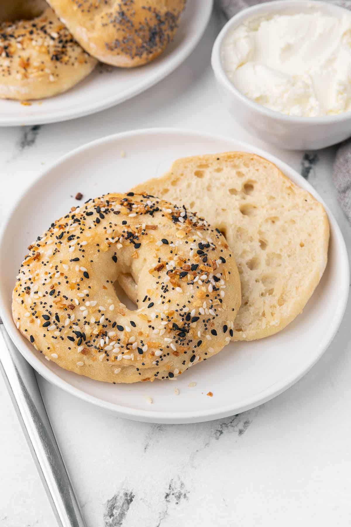 Bagel with everything but the bagel seasoning on a small plate spit in half with a small dish of cream cheese nearby.