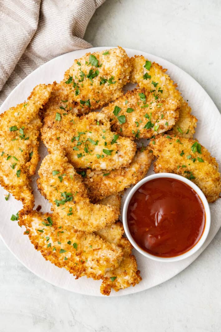 Air fried breadcrumb coated chicken tenders on a round plate garnished with fresh parsley with a small bowl of BBQ sauce for dipping.