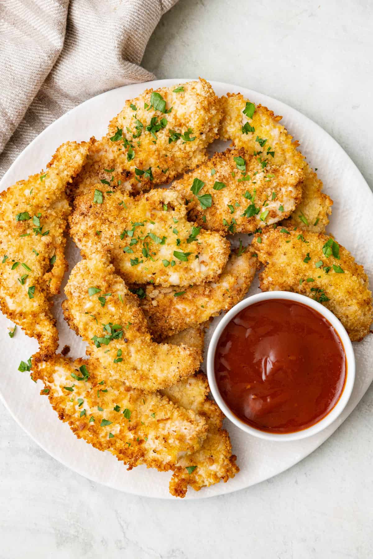 Air fried breadcrumb coated chicken tenders on a round plate garnished with fresh parsley with a small bowl of BBQ sauce for dipping.