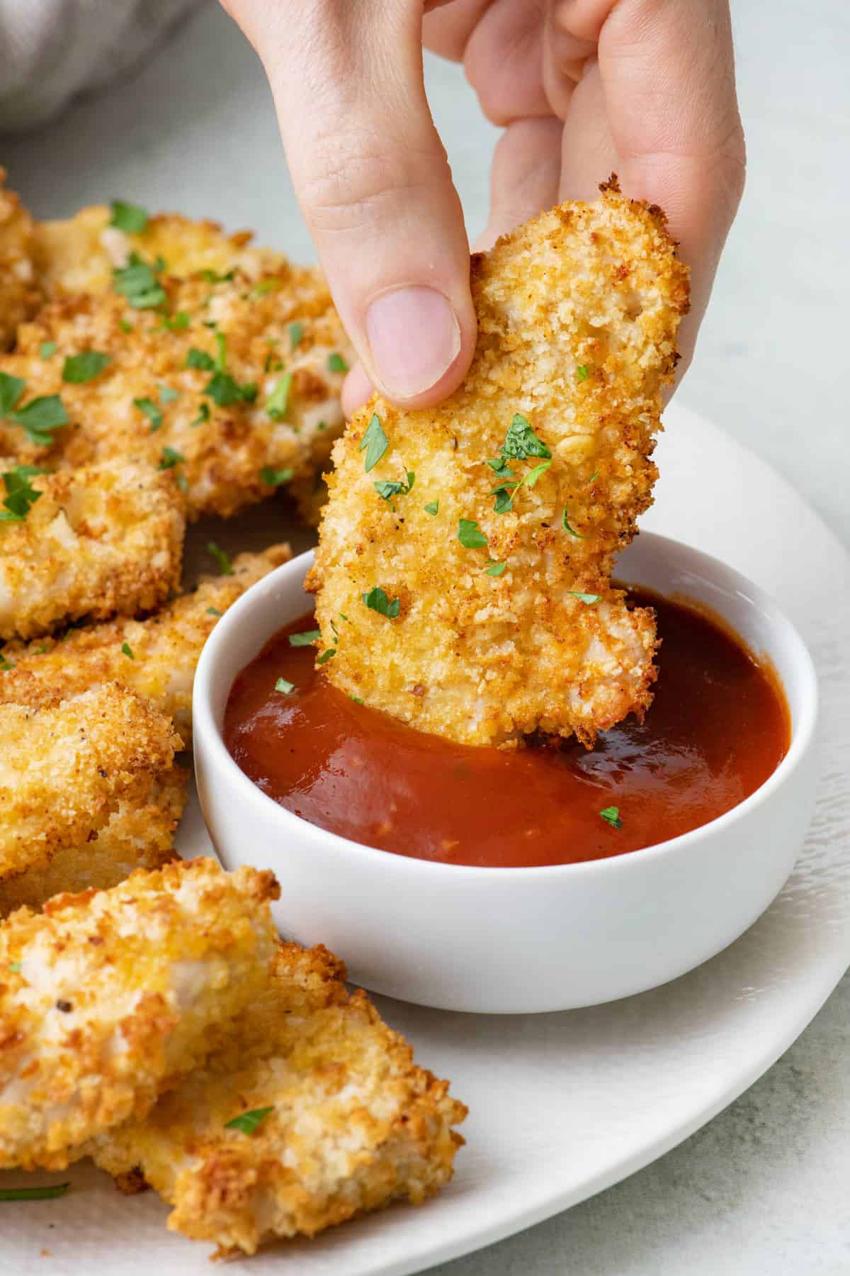 Hand holding an air fryer chicken tender and dipping it into BBQ sauce with more tenders around.