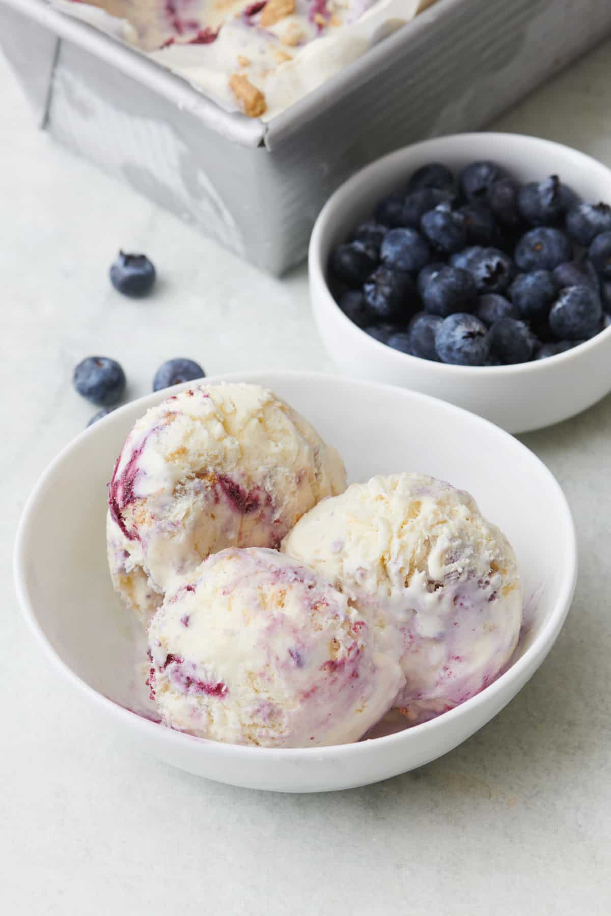 Blueberry ice cream in a bowl with fresh blueberries and the ice cream container nearby.