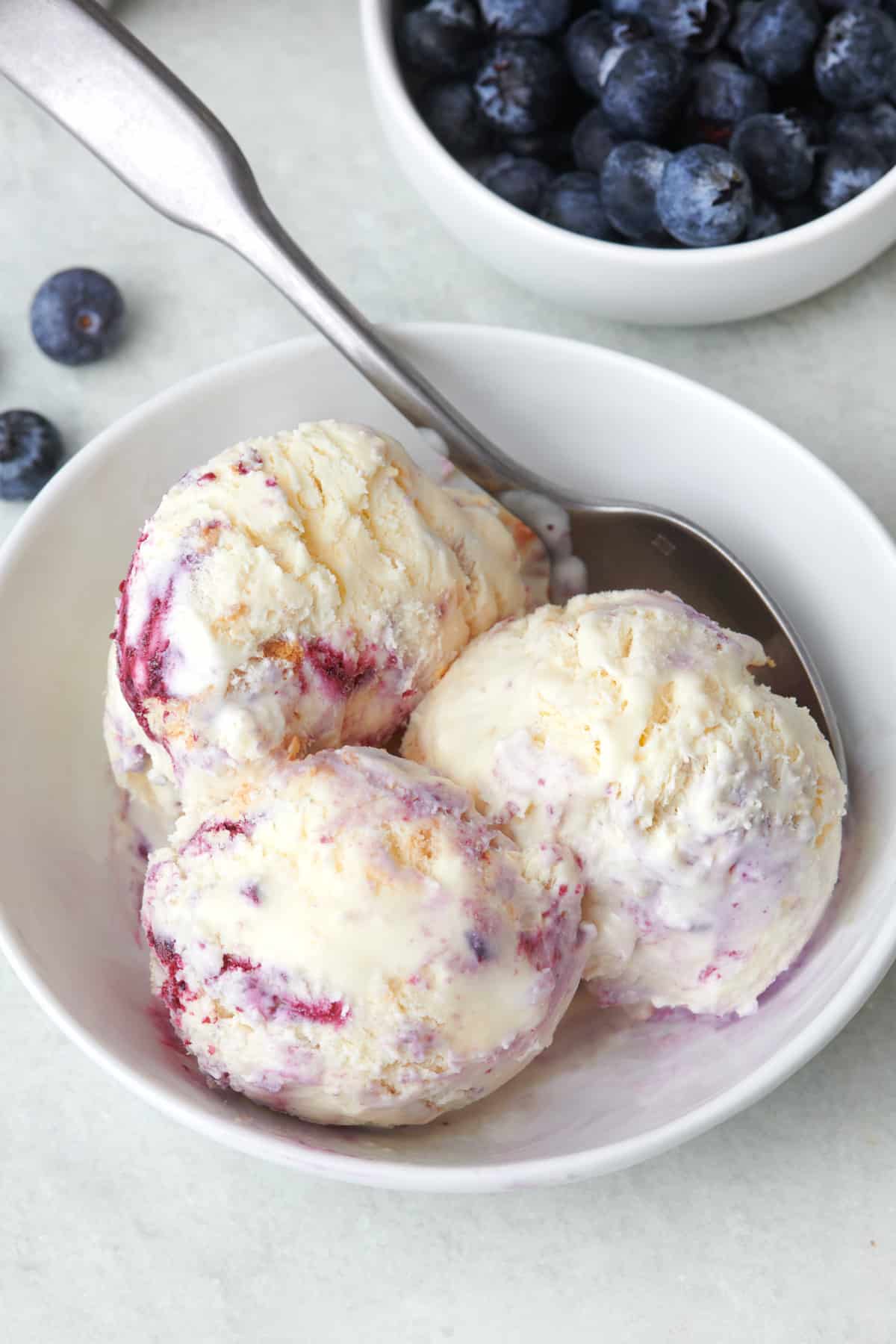 Three scoops of blueberry swirl ice cream in a bowl with a spoon dipped in and a fresh bowl of blueberries nearby.