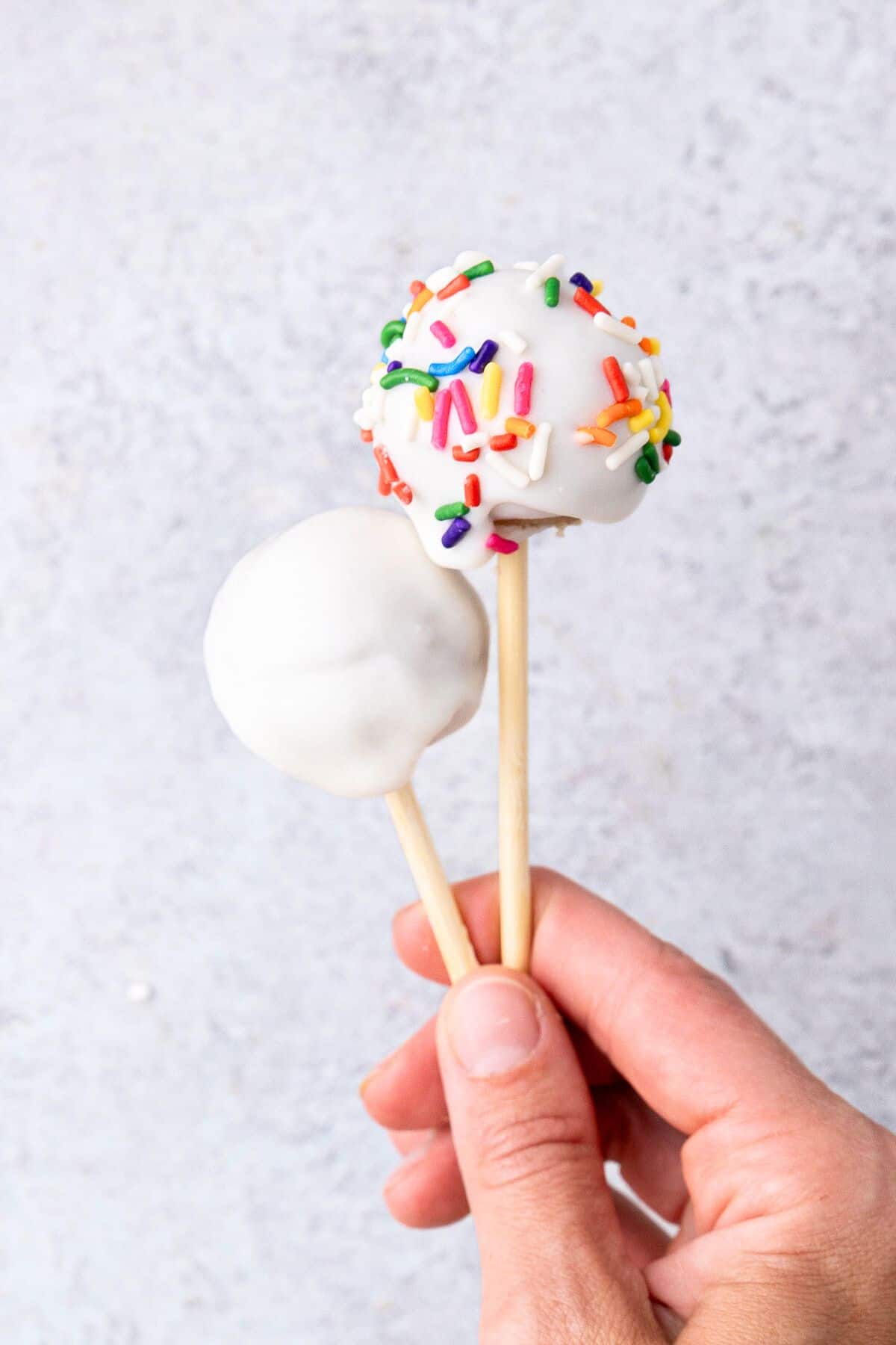 Hand holding 2 cake pops, one with sprinkles, one without.