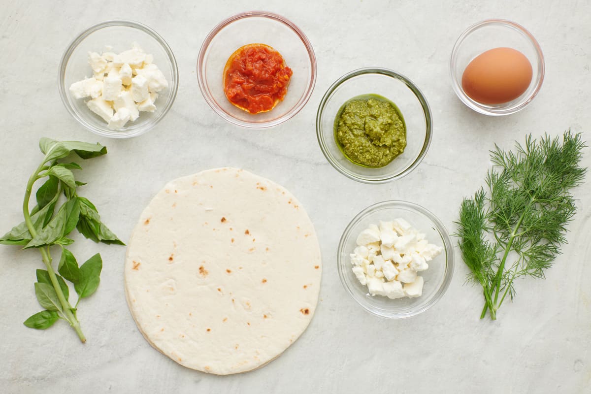 Ingredients for recipe: crumbled feta, harissa paste or pesto option, egg, dill, parsley, and tortilla shell.
