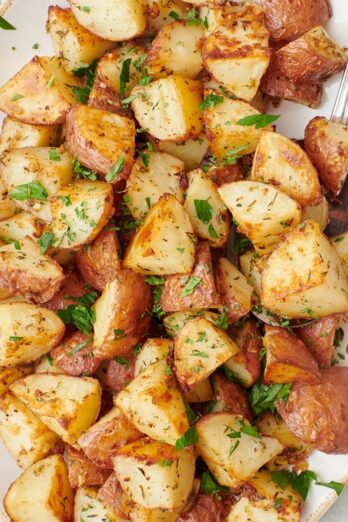 Square image of a long serving dish of garlic roasted potatoes garnished with fresh chopped parsley with a serving spoon dipped in.