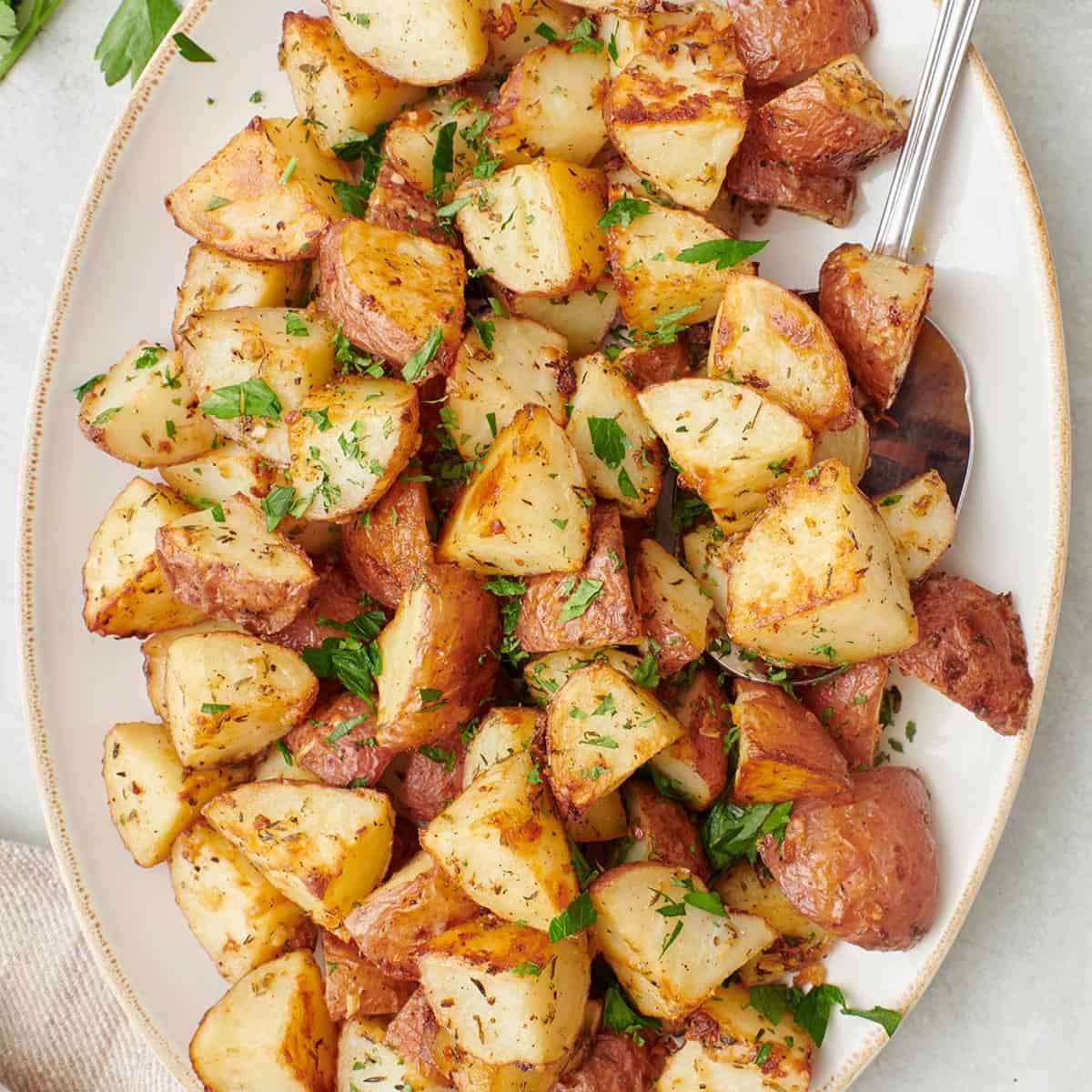 Square image of a long serving dish of garlic roasted potatoes garnished with fresh chopped parsley with a serving spoon dipped in.