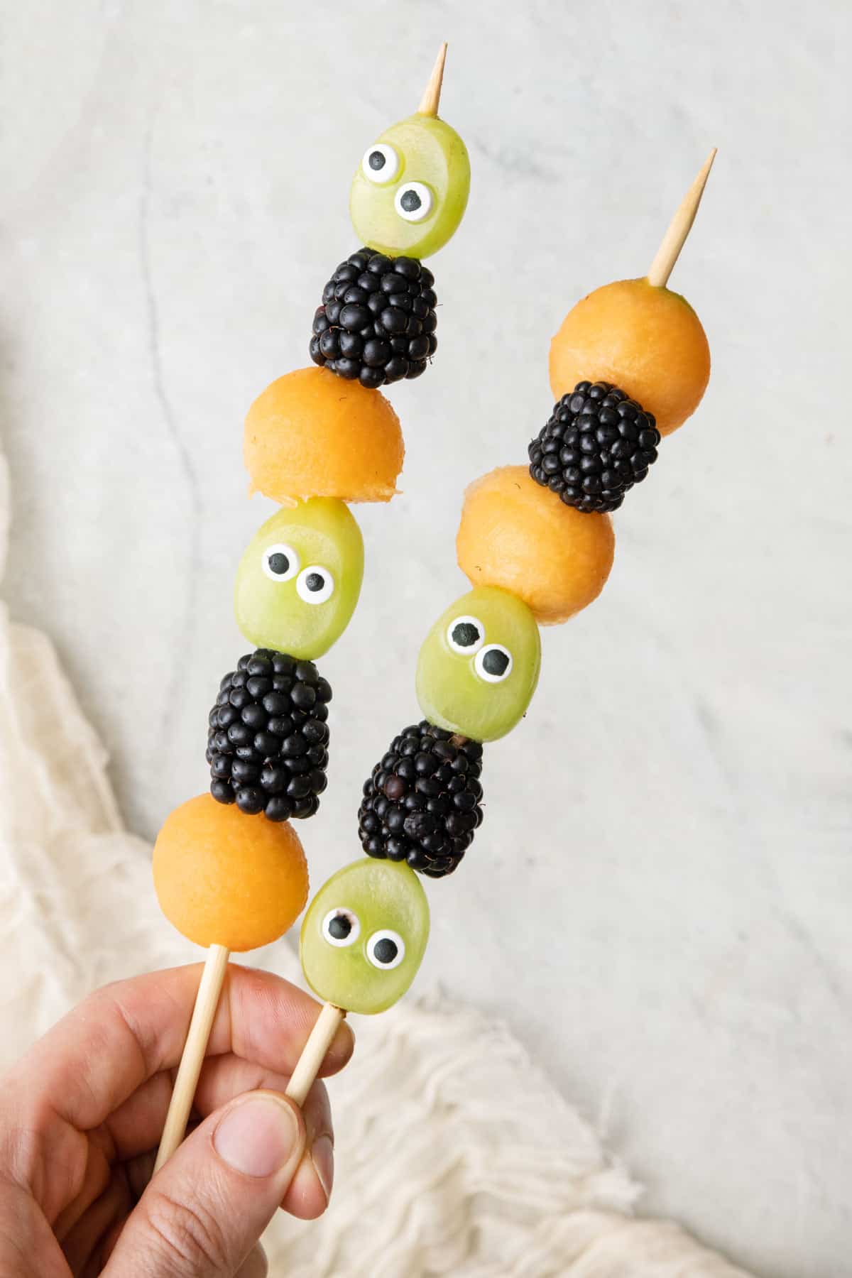 Hand holding two Halloween themed fruit kabobs with googly eyes.