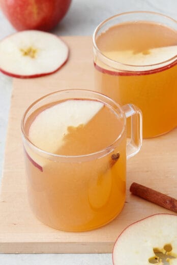 Two glass mugs of warm apple cider with an apple slice inside.