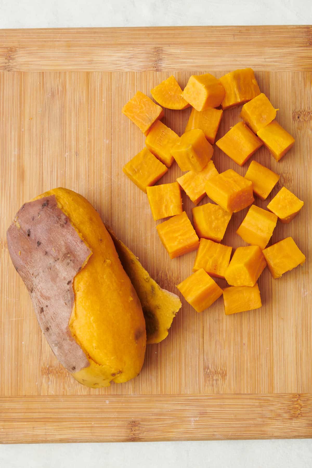 Two different boiled sweet potatoes on a cutting board: whole with the skin slightly pulled off and cubed.