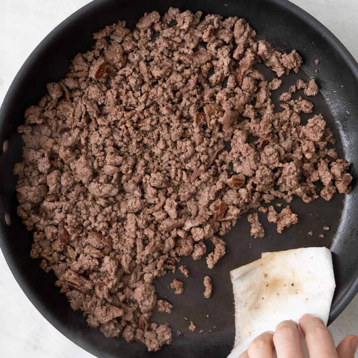 Hand holding folded paper towel dabbing extra fat from side of skillet.