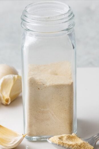 Garlic powder made from garlic cloves in a spice jar with more cloves nearby.