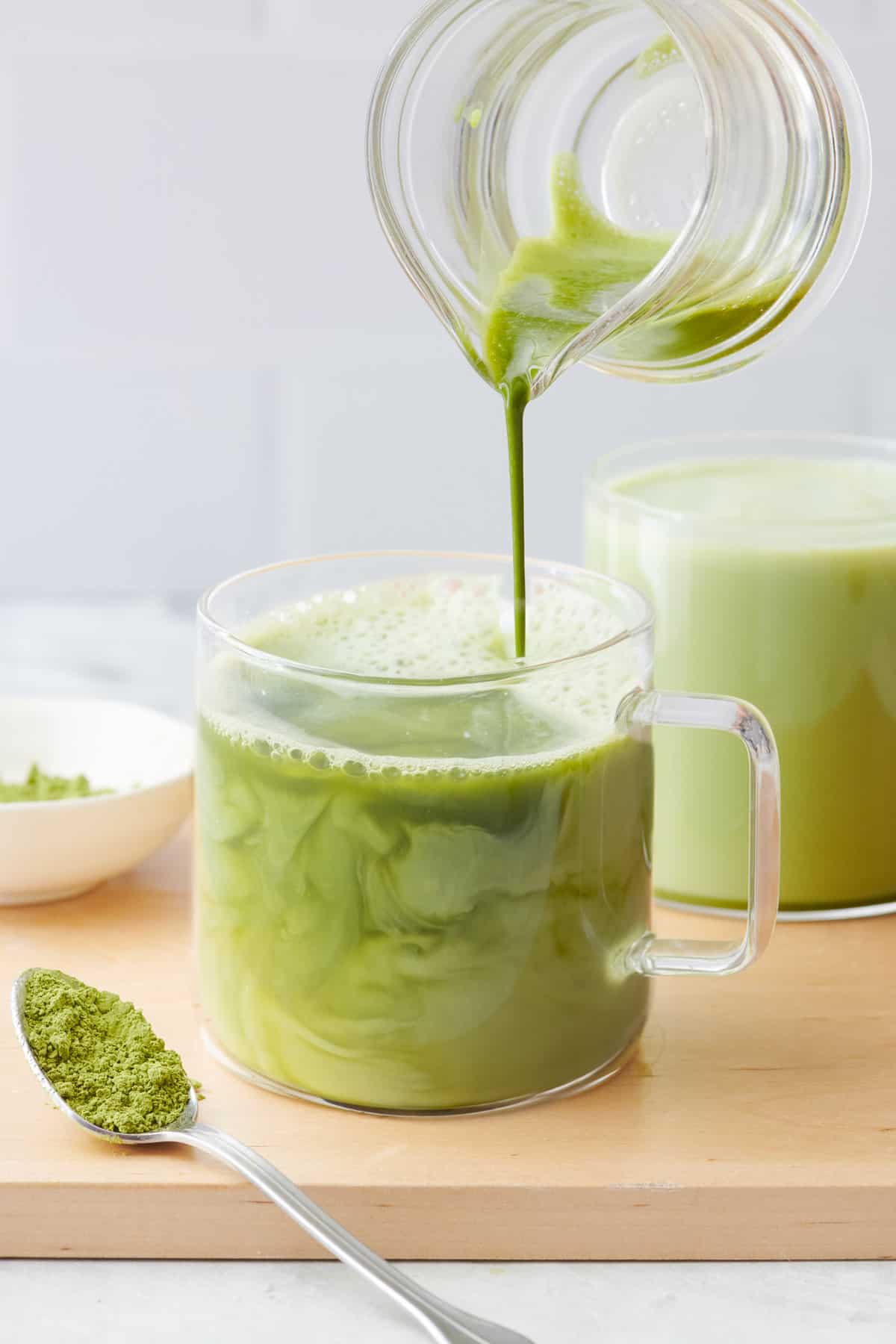 Matcha latte mixture is poured in to warm milk in a glass mug with another mug of latte nearby.